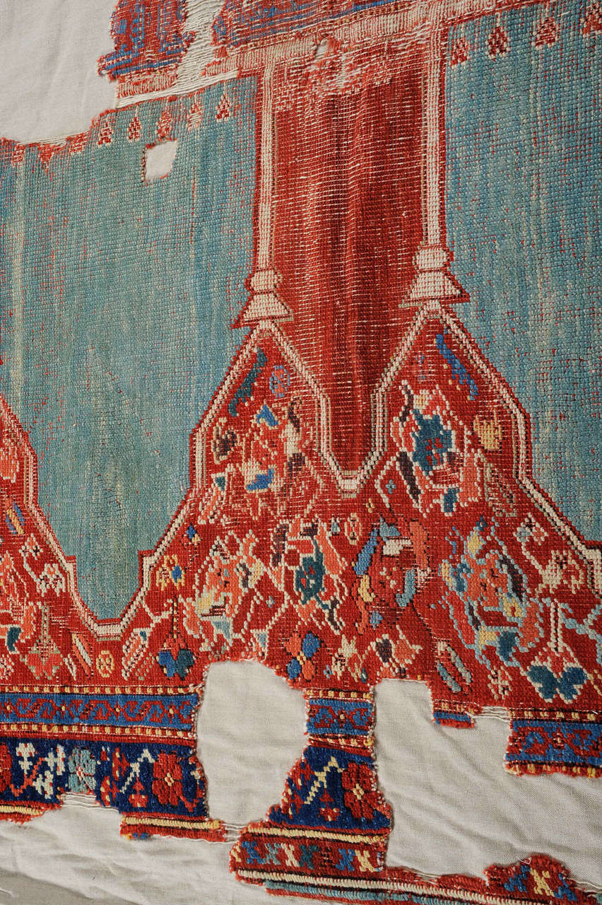 Un fragmento de alfombra otomana verdaderamente majestuoso, decorado con una secuencia de cinco nichos con capiteles que desembocan en un fondo verde agua. Montado profesionalmente sobre un soporte de algodón, este monumental tejido arquitectónico